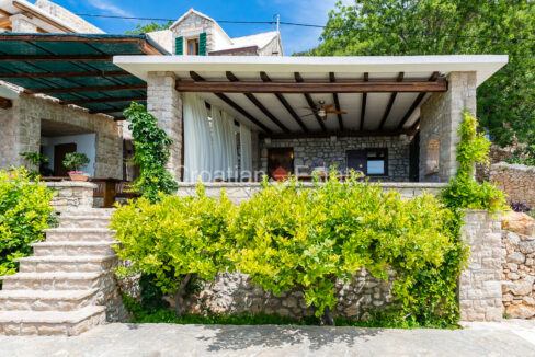 An autochthonous house for sale on Brac, Croatia, with a stone facade, a covered terrace, stairs, and plants.