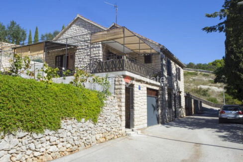 A stone house for sale on Brac, Croatia, with terraces, a stone fence with a plant on it, and an asphalt road next to it.