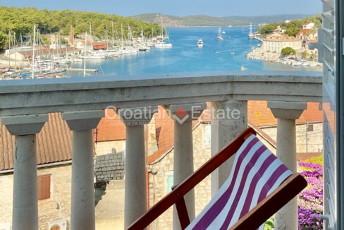 A house for sale on Brac, Croatia, with a balcony with a sun lounger, a fence, and a view of the sea, vegetation, and houses.