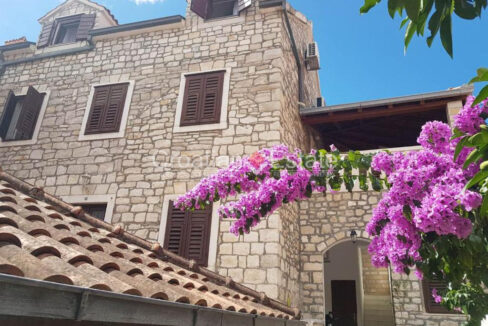 A stone house for sale on Brac, Croatia, with a stone facade, brown shutters, and a roof and flowers in front of it.