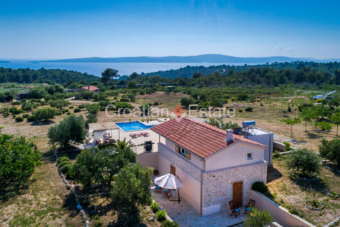 A villa with a partially stone facade, a clay roof, and a patio with a pool and garden with plants, a land, and the sea.