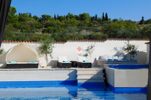A patio of a villa for sale on Brac, Croatia, with a pool, a sunbathing area, and lush nature behind its fence.