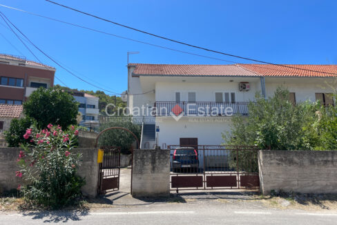 A house for sale on Ciovo, Croatia, with a balcony and a yard with a stone fence and metal gates and a parking area inside.