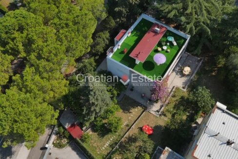 A villa for sale in Split, Croatia, from above, with a roof terrace, lush vegetation around it, and a house on the right.