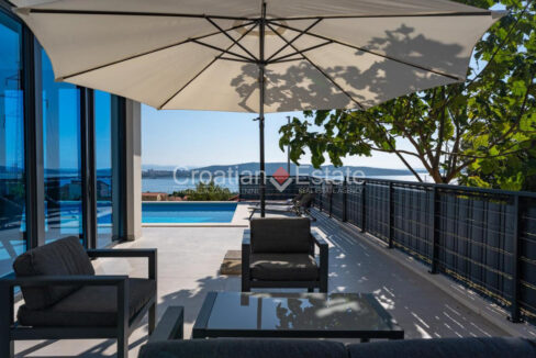 A villa for sale near Split, Croatia, with a terrace a table, armchairs, a sun umbrella, a fence, a pool, and a sea view.