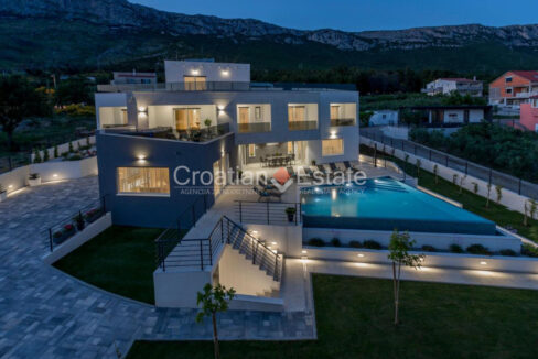 An illuminated villa for sale in Split, Croatia, with a patio with a terrace with a pool, grass, and an empty paved area.