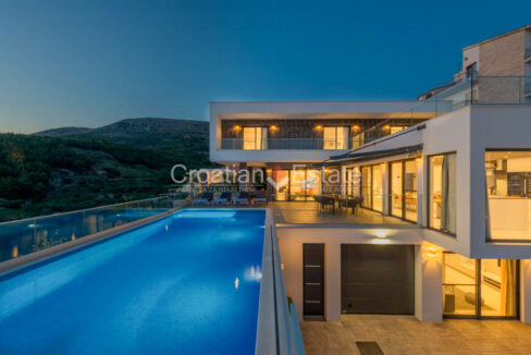 An illuminated villa for sale near Split, Croatia, at dusk, with a balconies and terraces, one of which has a pool.