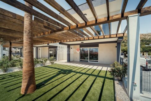 A house for sale in Trogir, Croatia, with a yard with lawn, a tree, pebbles, plants, and a pergola.