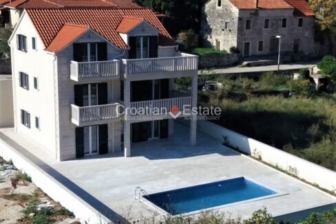 A stone villa for sale on Brac, Croatia, with balconies, and a fenced terrace with a pool and a section with plants.