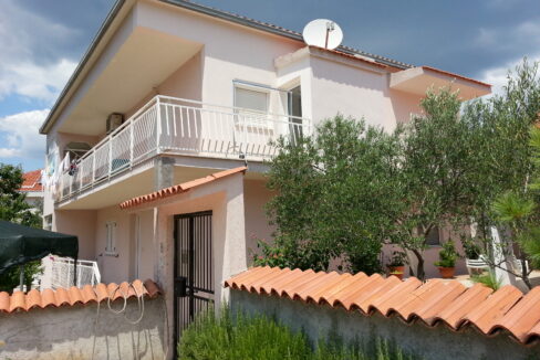 A house for sale on Ciovo, Croatia, with a balcony, a terrace, trees, plants, and a fence with a metal door with bars.