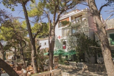 A hotel for sale in Makarska, Croatia, in an area with houses, a promenade, trees, and a terrace of a restaurant or a café.