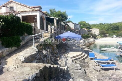 A seafront, stone house for sale on Vis, Croatia, with a terrace, a walkway with stairs, a beach, and the sea with boats.